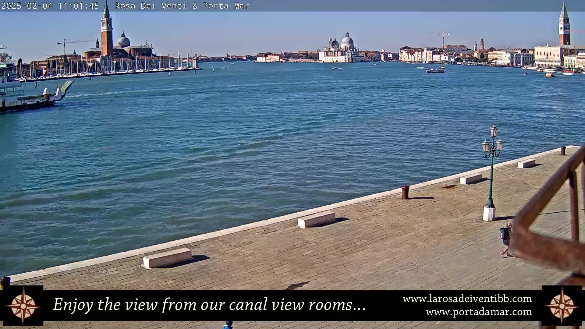 A sunny day in Venice shows a waterfront paved area with a lamppost and a person walking, overlooking a canal with boats and buildings, including a large church with domes and a tall bell tower, in the distance.