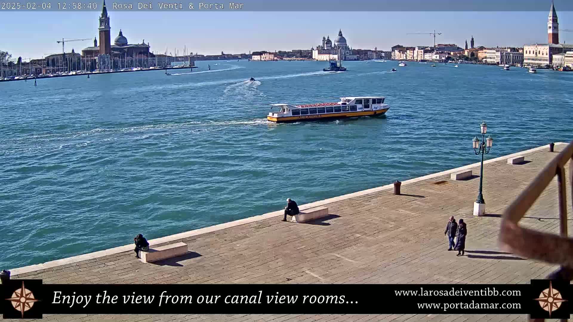 A waterfront paved area with a few people sitting and walking, overlooking a canal with a boat and a distant city under a sunny sky.