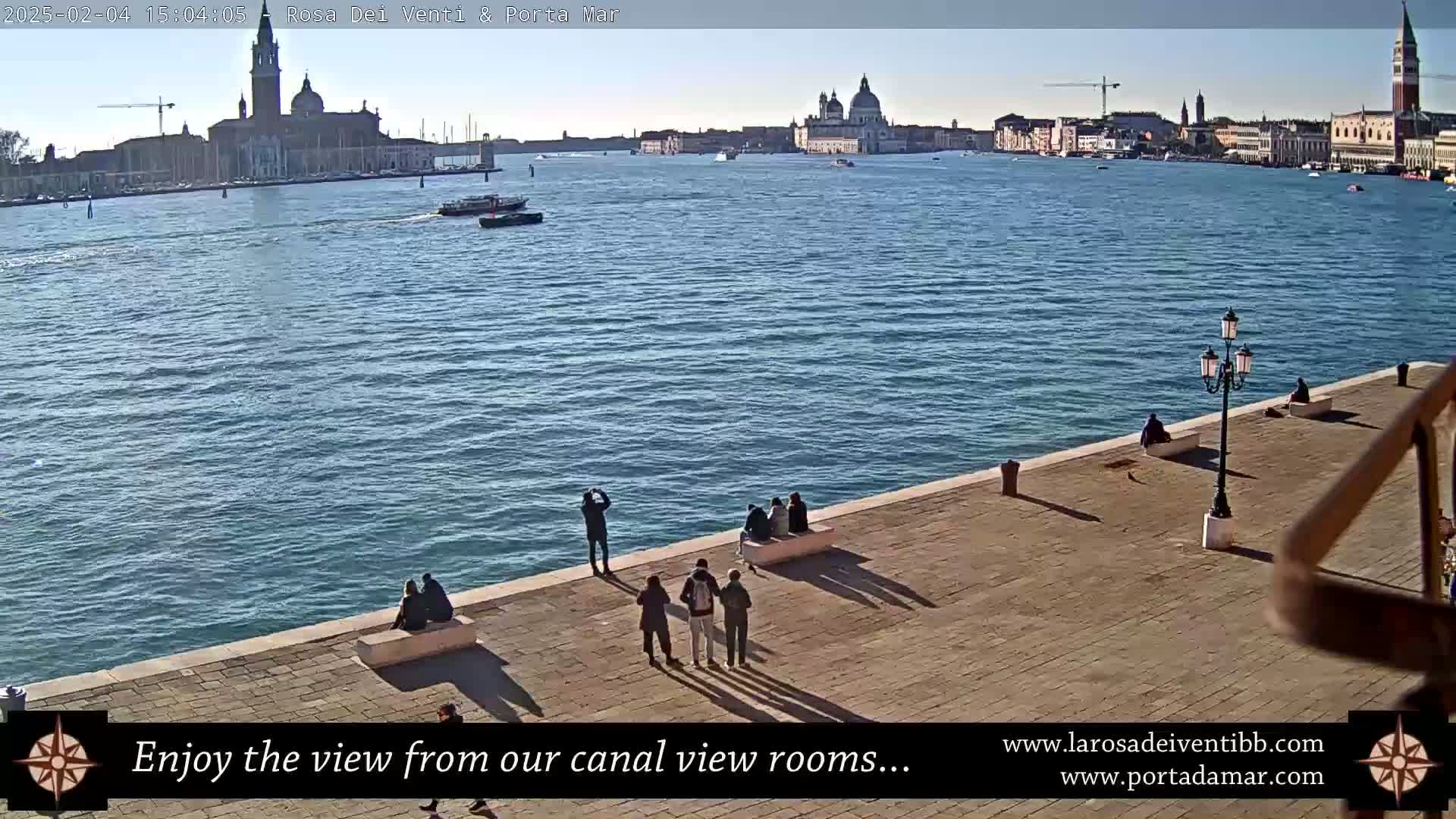 A calm, sunny waterfront scene shows several people relaxing on a paved area overlooking a canal, with distant Venetian buildings visible across the water.