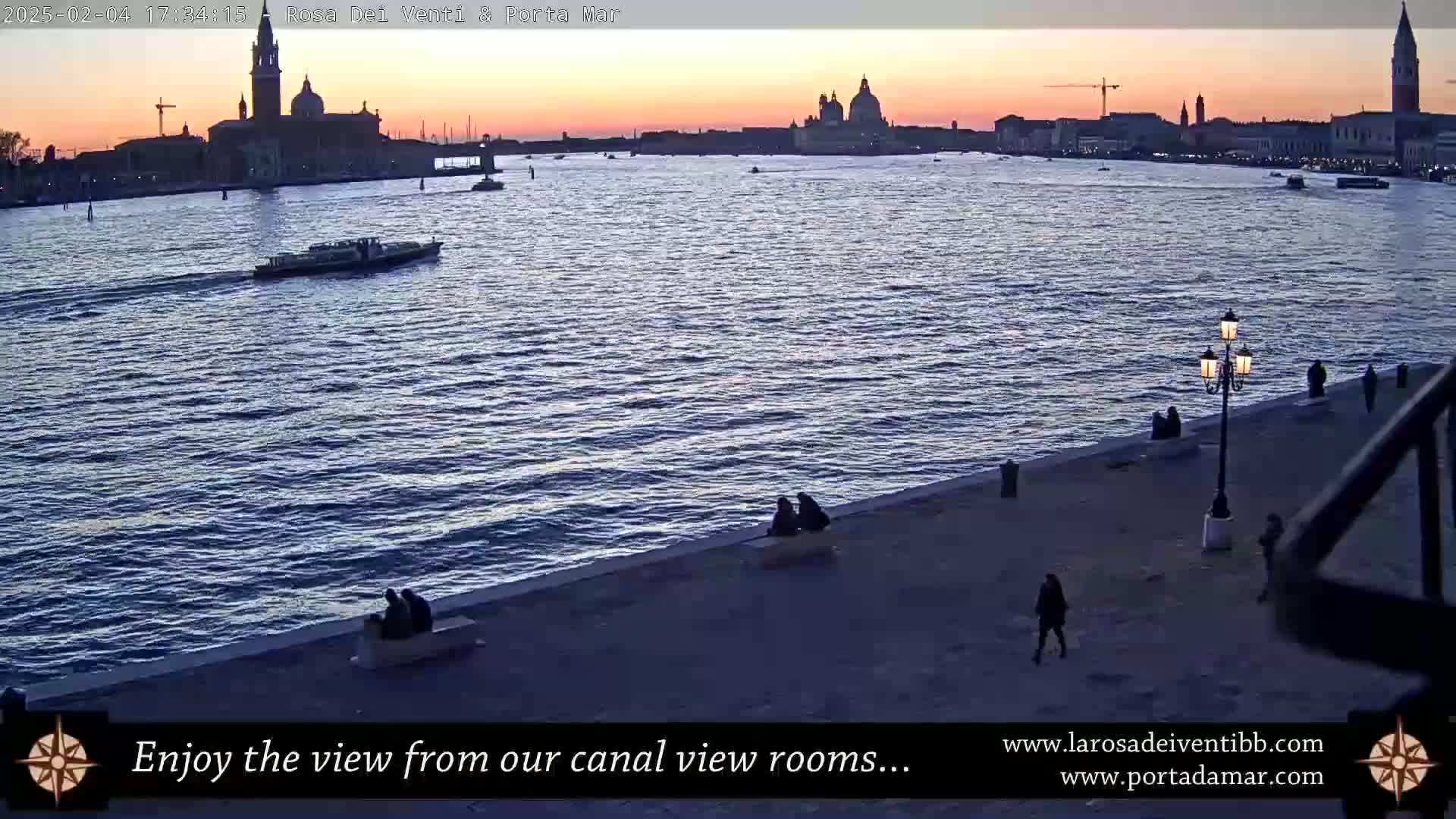 A calm body of water at sunset shows a city skyline with several prominent buildings, a few boats, and several people walking along a stone embankment.