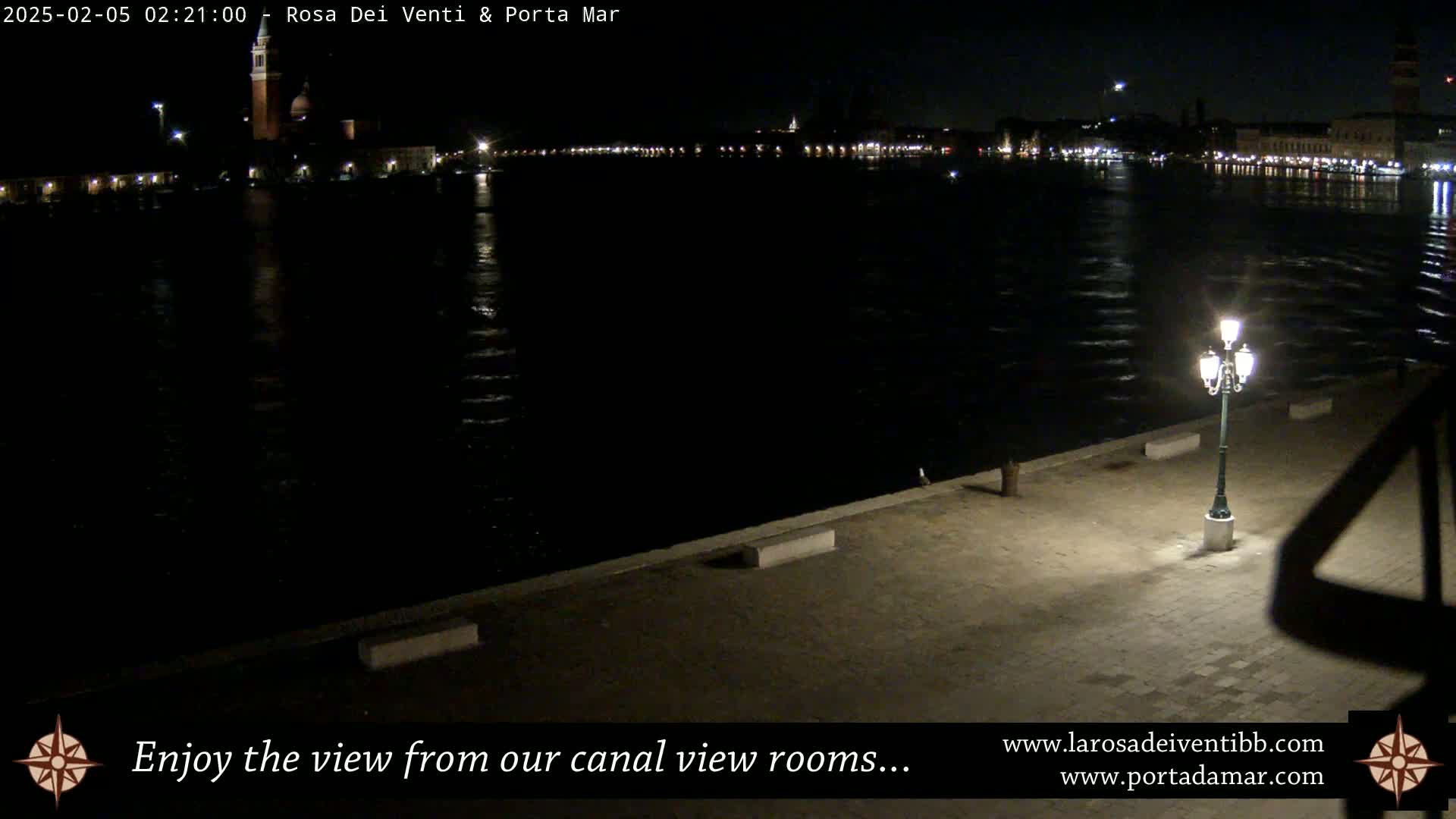 A nighttime view across a dark canal shows a distant illuminated cityscape and a single lit lamppost on a paved waterfront.