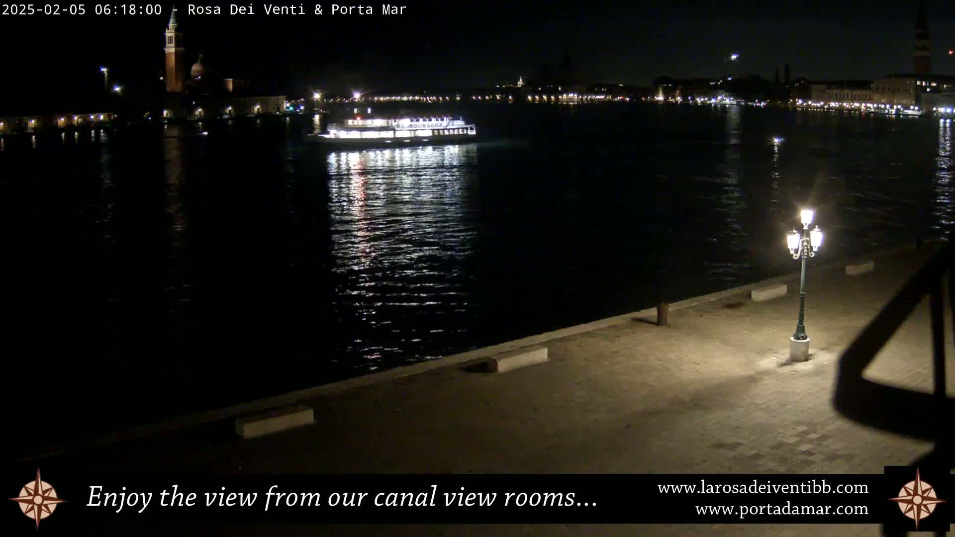A nighttime view of a waterway shows a boat on the water, city lights in the distance, and a lit lamppost on a paved waterfront.