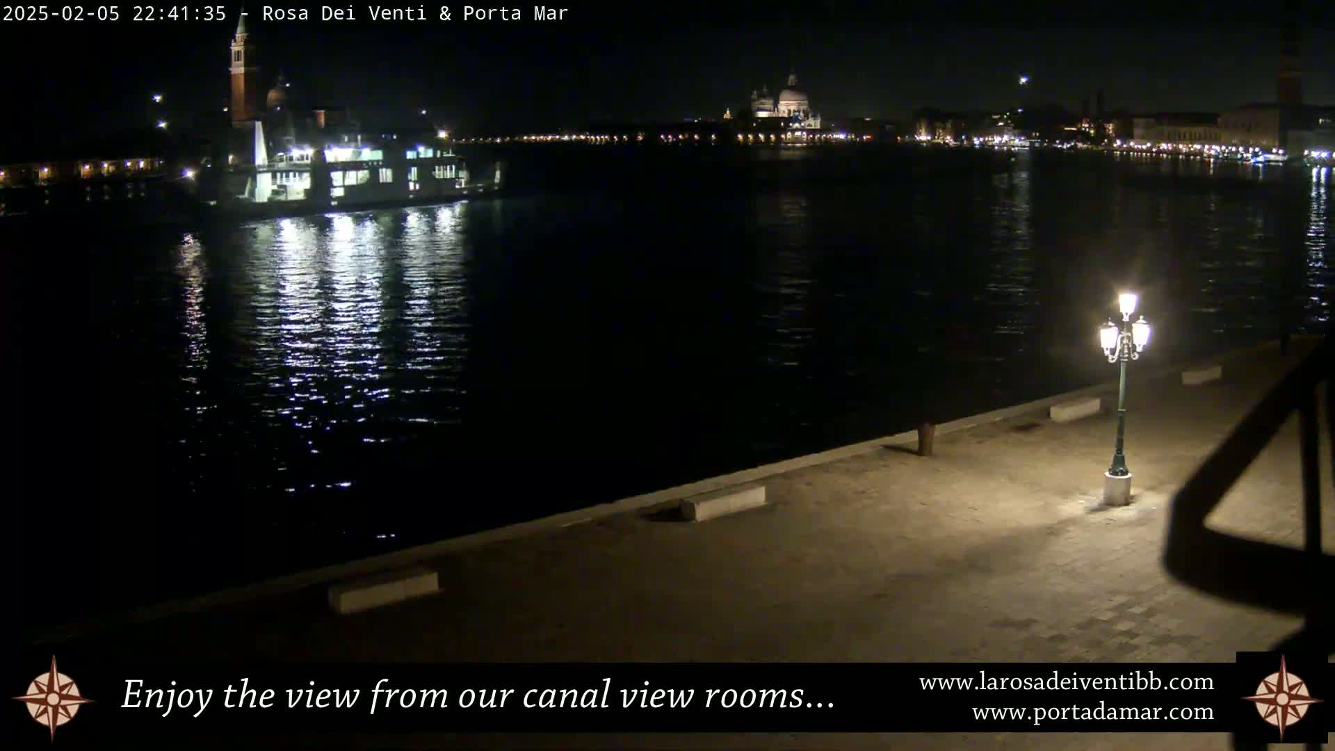 A nighttime view across a calm canal shows a ferry and the illuminated cityscape beyond, a single lamppost standing in the foreground.