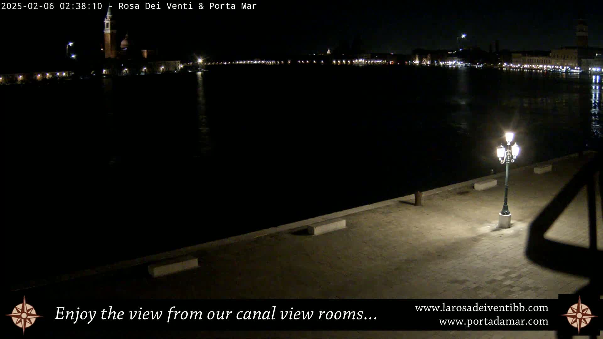 A nighttime view of a canal, with a city skyline faintly visible across the water, illuminated by a single lamppost on a stone embankment.
