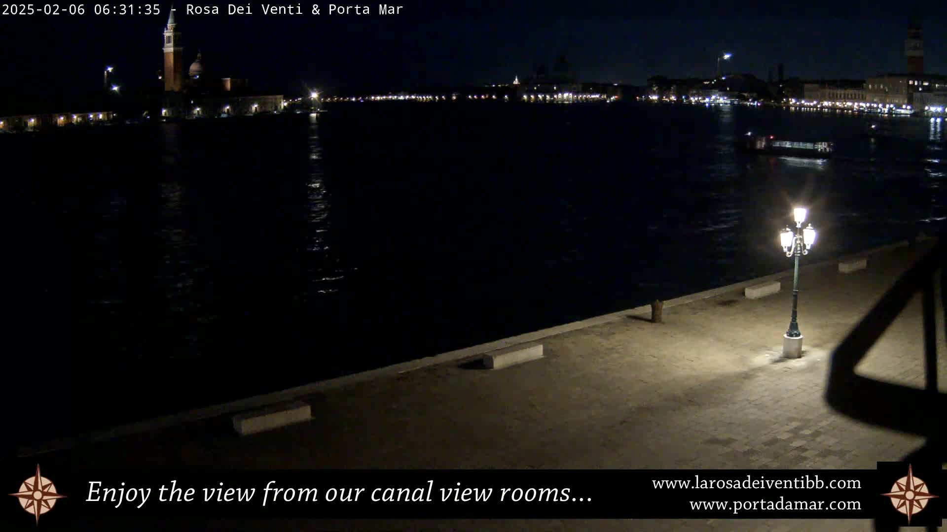 A nighttime view of a dark canal reflecting city lights, with a single illuminated lamppost on a paved waterfront.