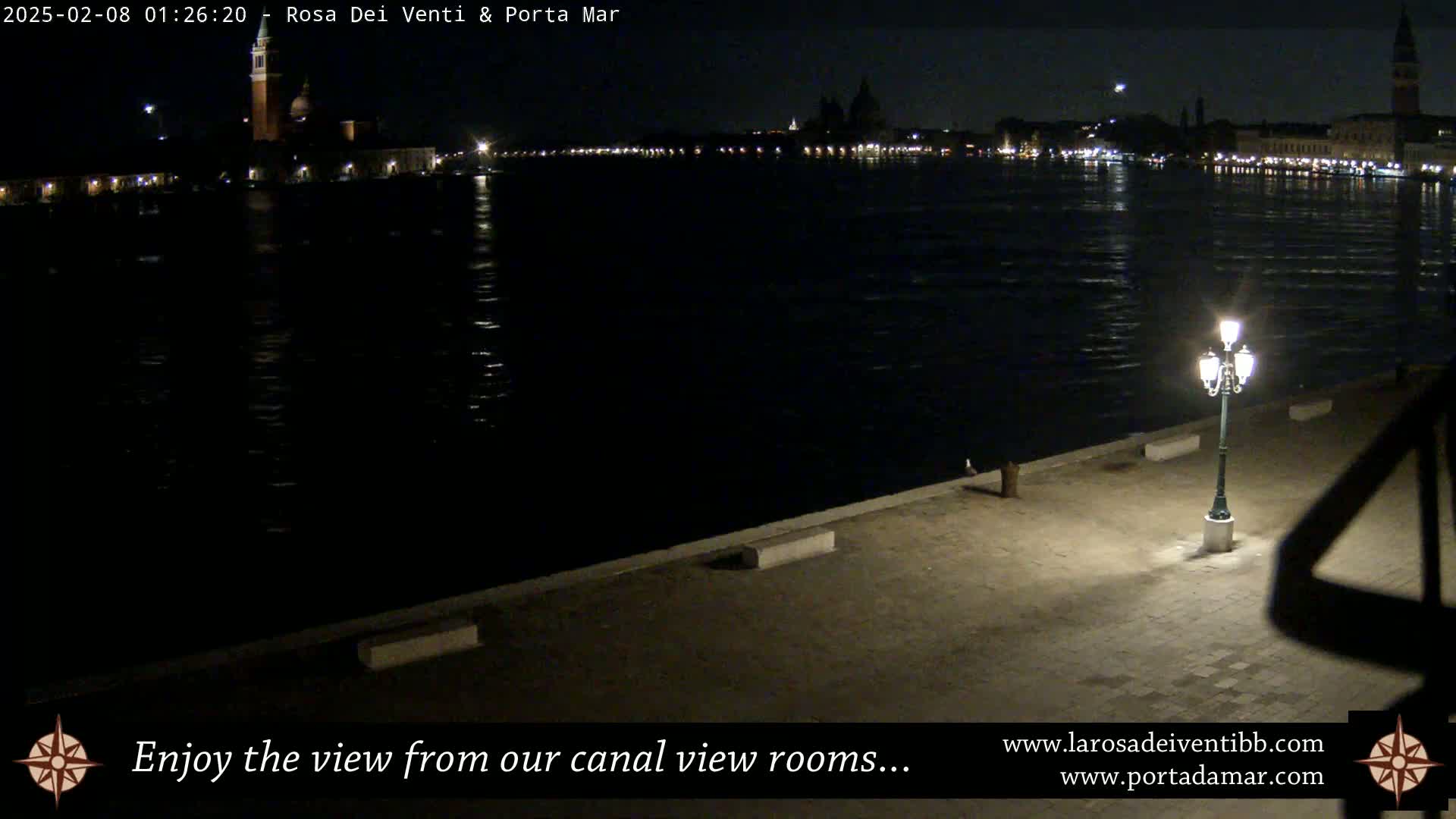 A nighttime view across a calm canal shows a city skyline with illuminated buildings, and a single lit lamppost in the foreground.