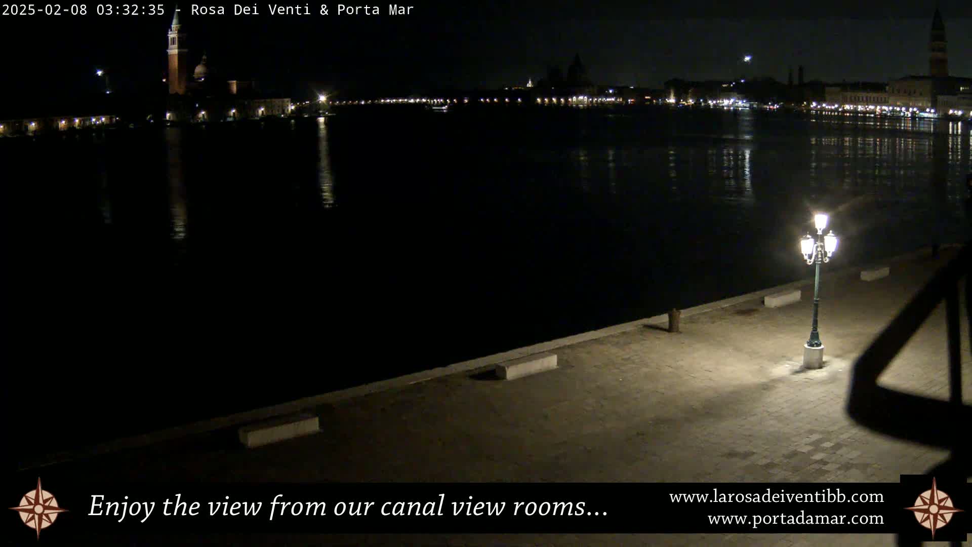 A nighttime view of a calm canal reflecting the lights of a city across the water, a single lit lamppost standing on a paved walkway in the foreground.