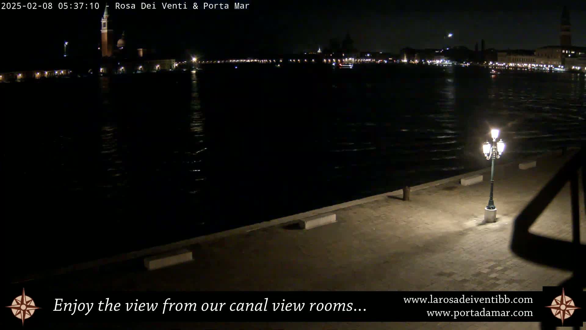 A nighttime view across a dark canal shows the illuminated cityscape in the distance, with a single lit lamppost on a paved waterfront in the foreground.