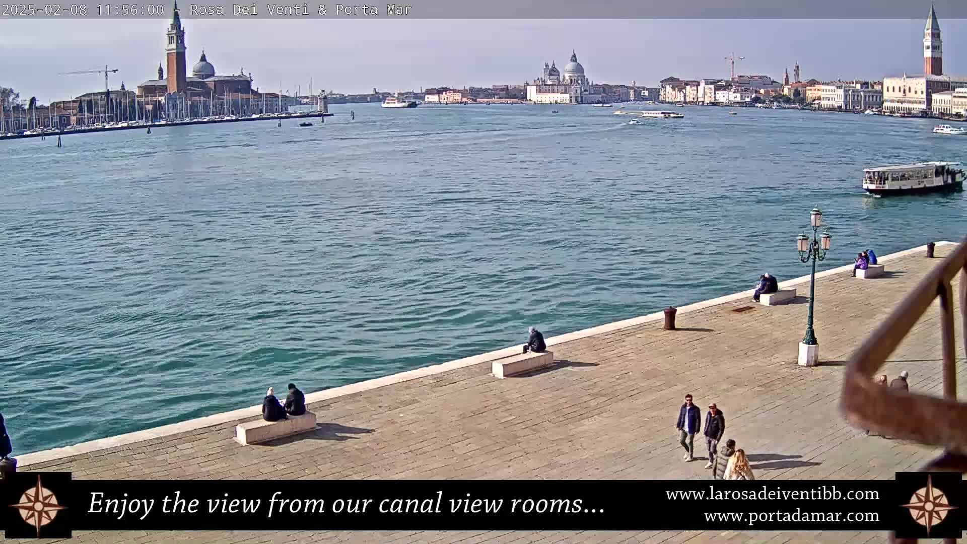 A sunny day in Venice shows a calm canal with several people sitting and walking along a stone embankment, overlooking a cityscape of buildings and churches.