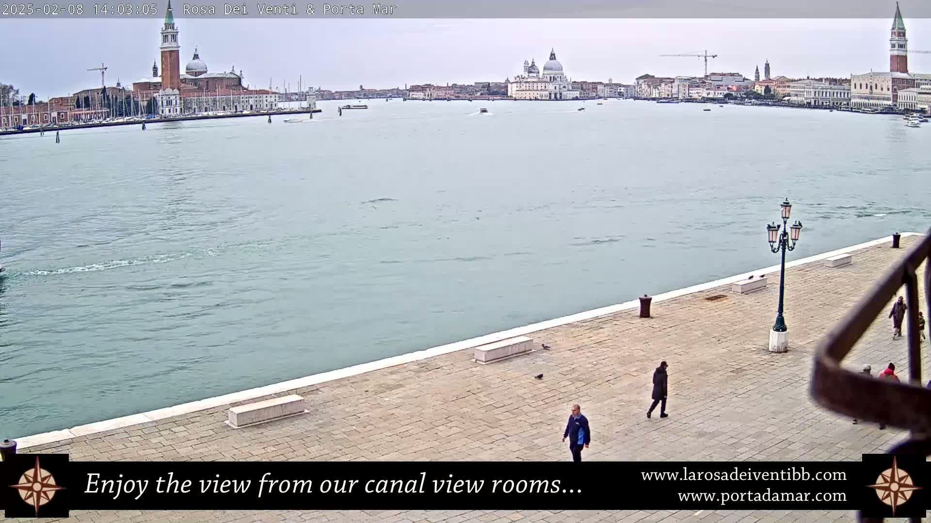 A wide shot of a calm canal in Venice, Italy, on an overcast day, showing a paved walkway with several people and a view of buildings across the water.