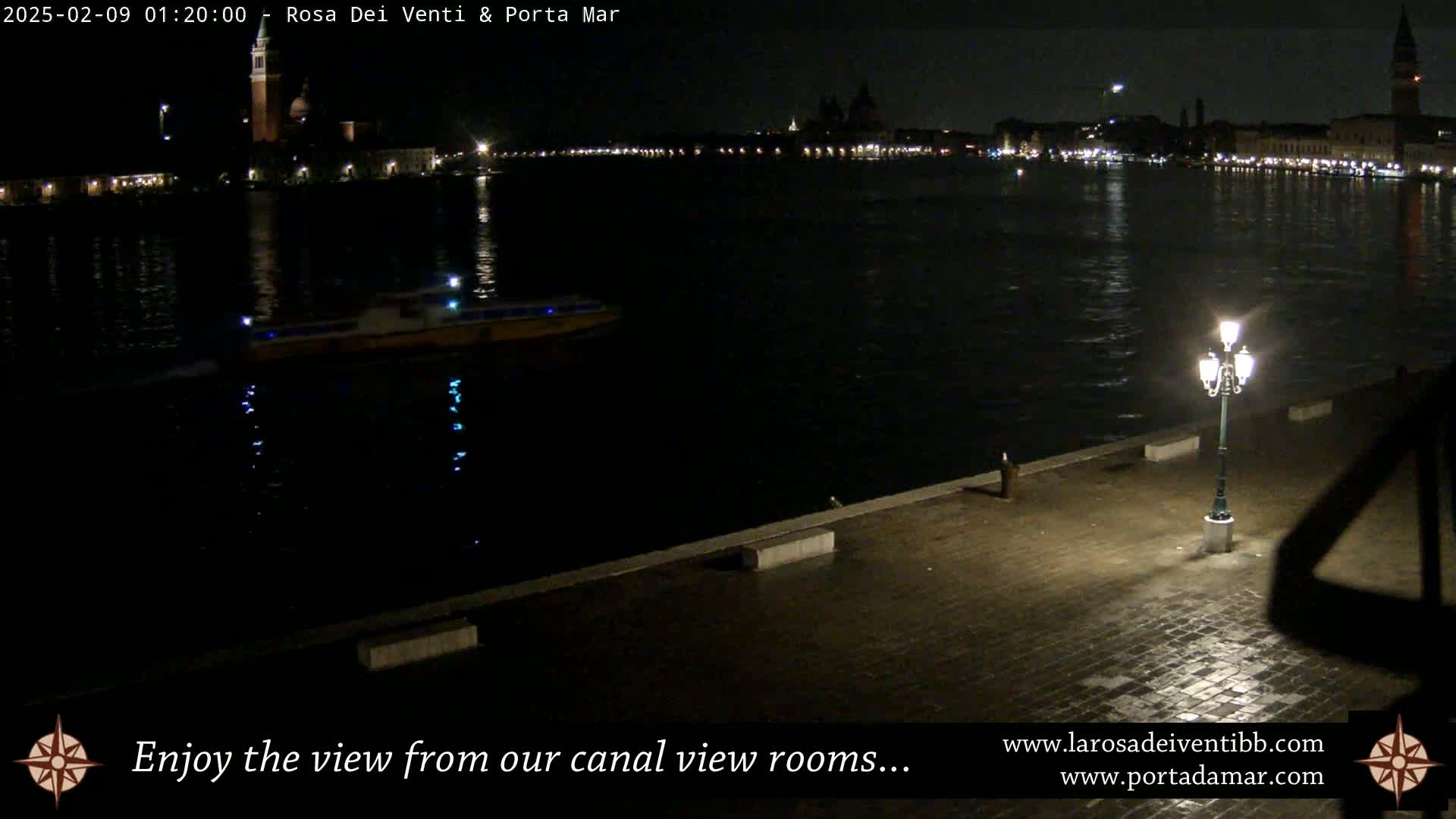 A nighttime view across a dark canal shows a distant cityscape with lights, a boat on the water, and a single lit lamppost on a paved waterfront.