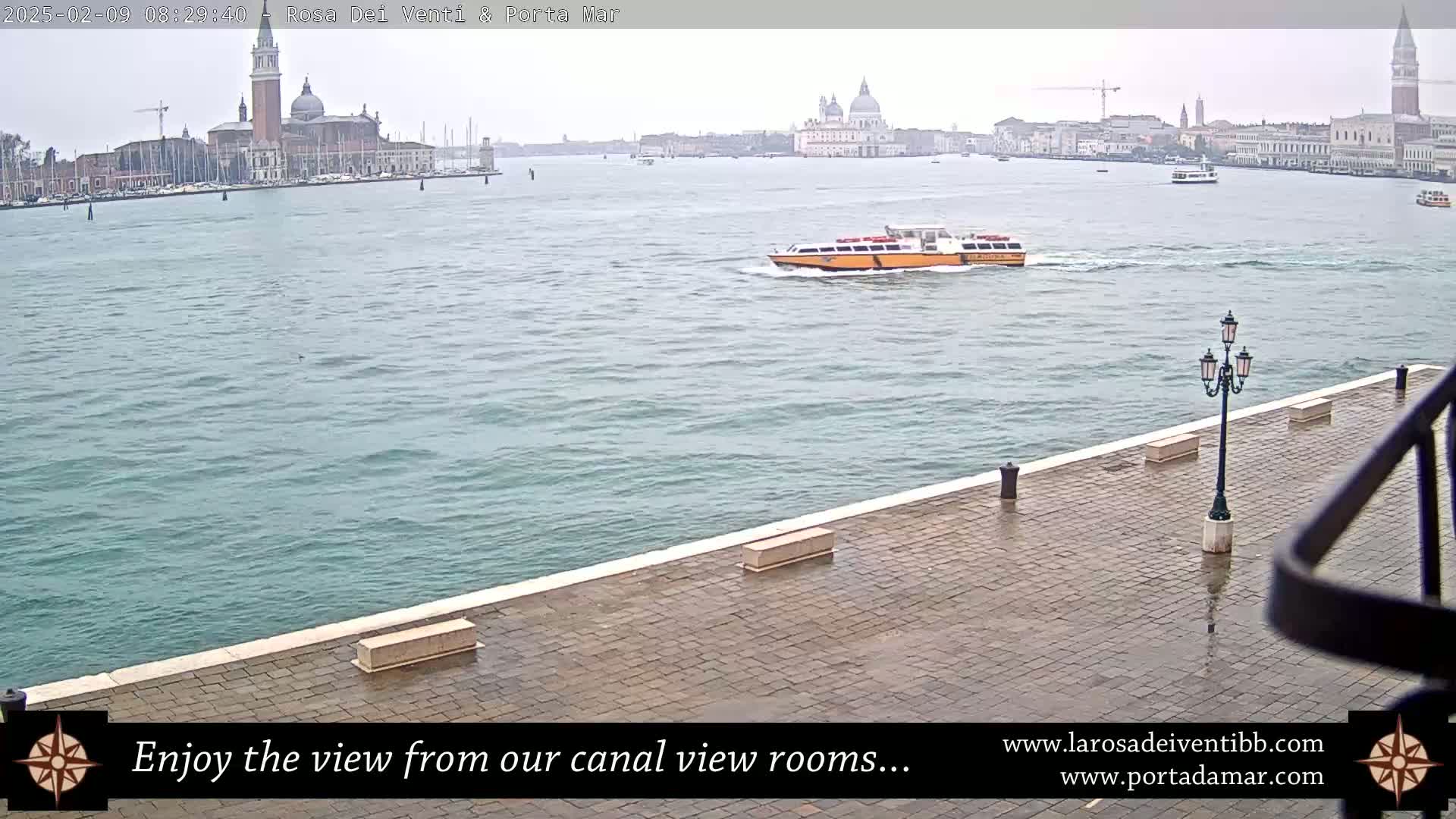 A waterway view shows a paved area with a lamppost, a yellow water taxi, and a distant cityscape under an overcast sky.