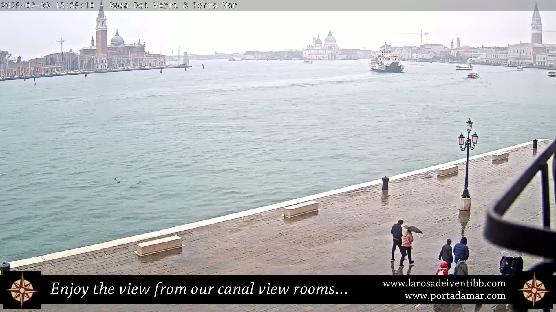 A rainy view of a canal in Venice shows several people walking along a wet paved walkway toward a distant cityscape with boats and church domes.