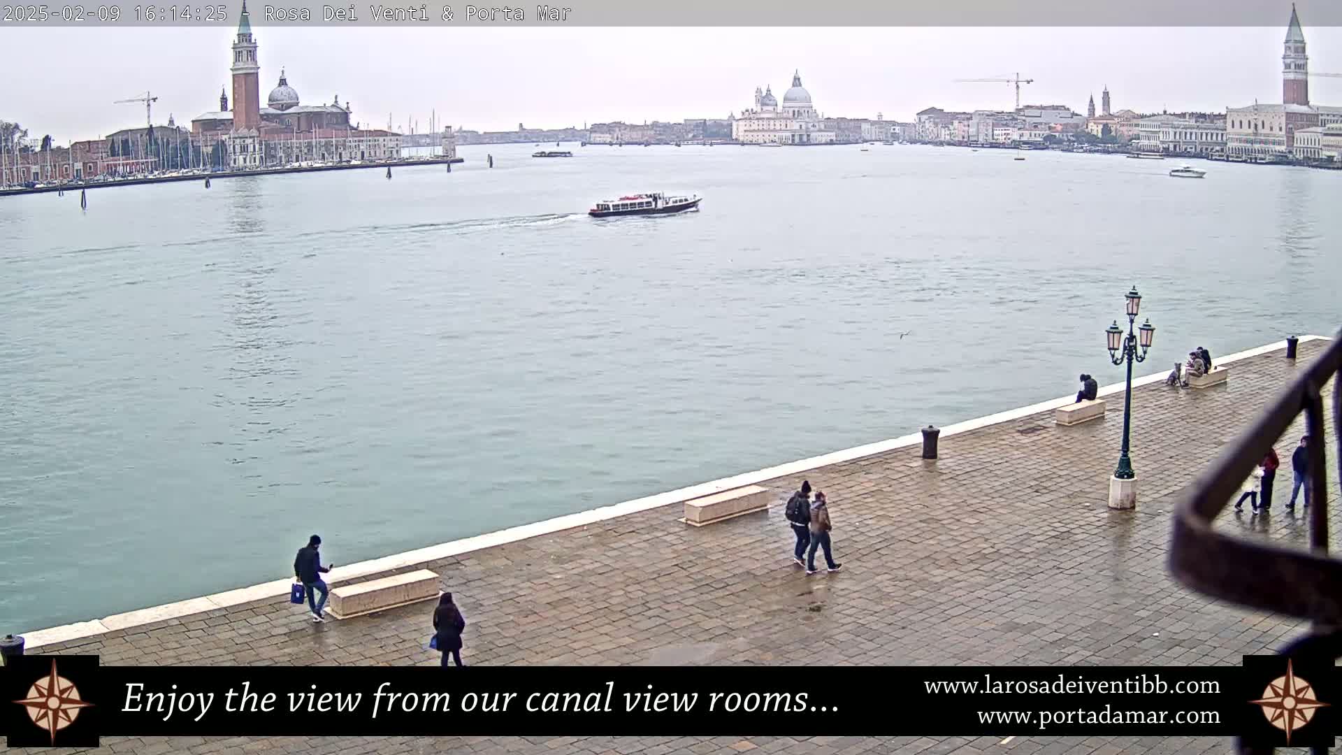 A wide shot of a waterway in Venice, Italy, on an overcast day, shows a paved walkway with several people walking, sitting, and standing, overlooking the water where a boat is visible and the city's buildings are in the distance.