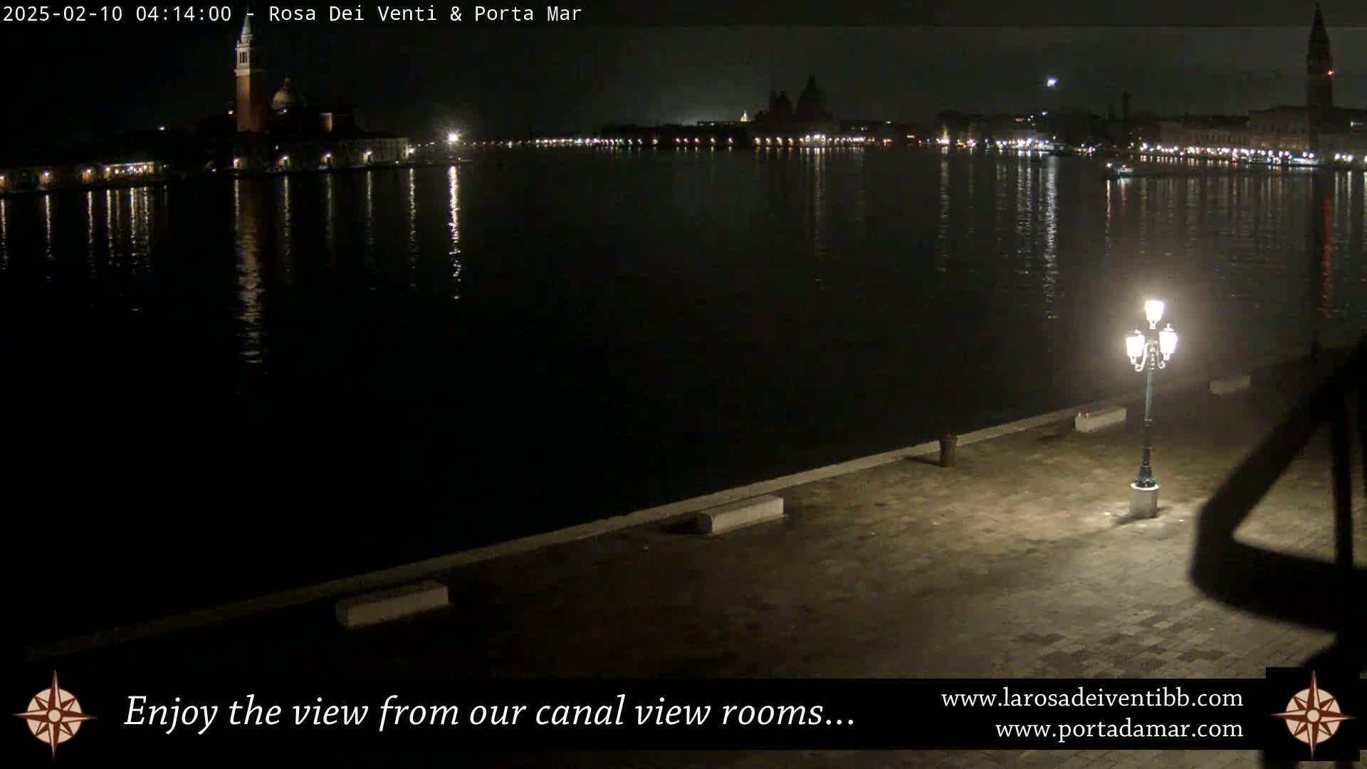 A nighttime view across a calm canal shows a cityscape with lights reflecting on the water, illuminated by a single lamppost on a stone embankment.