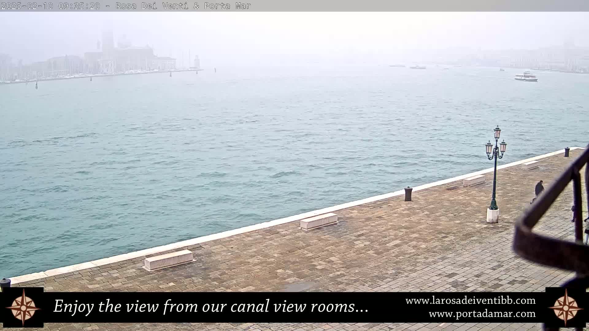 A hazy view of a waterway shows a city in the distance, with a paved walkway and lamppost in the foreground.
