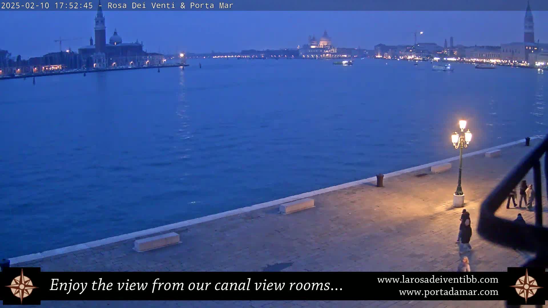 A twilight view of a calm canal in Venice, showing a waterfront walkway with several people walking, and buildings and churches across the water.