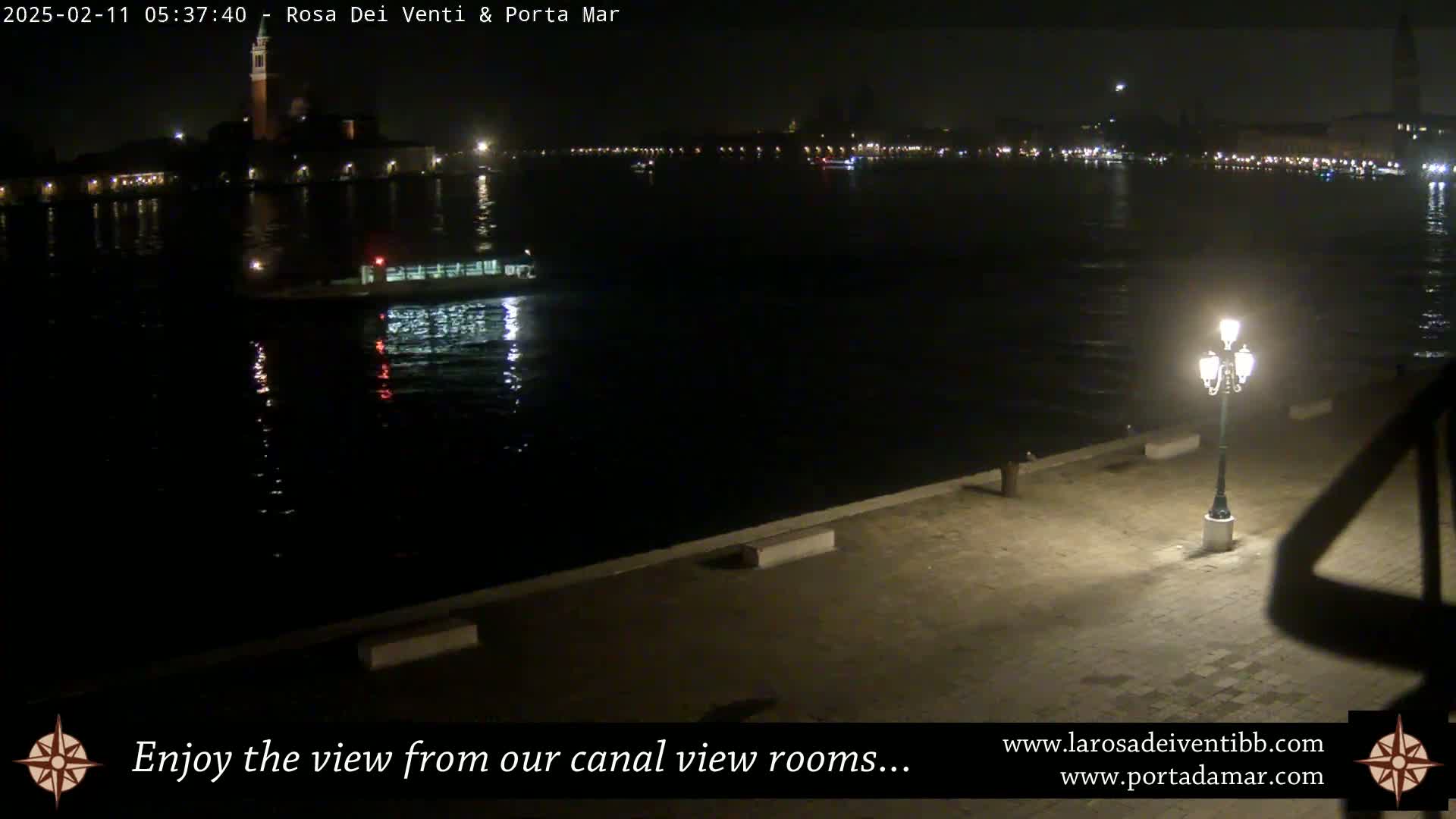 A nighttime view of a canal, showing a boat on the water, city lights in the distance, and a lit lamppost on a paved waterfront.