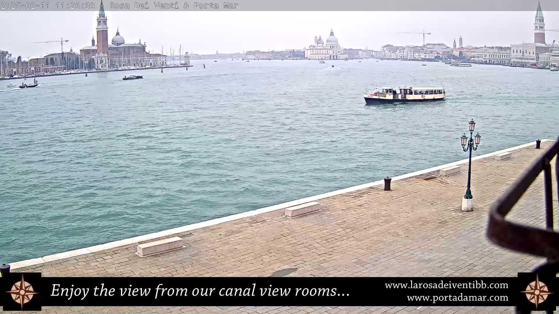 A waterway view shows a paved waterfront with a lamppost, a boat, and a distant cityscape under an overcast sky.