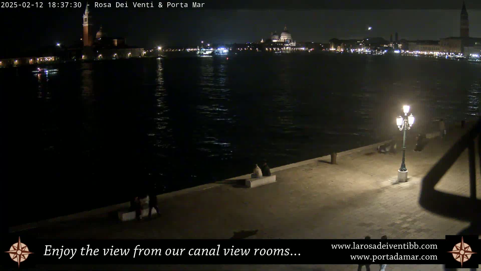 A nighttime view across a dark canal shows illuminated buildings on the far shore and a few people sitting on a waterfront embankment under a lit lamppost.