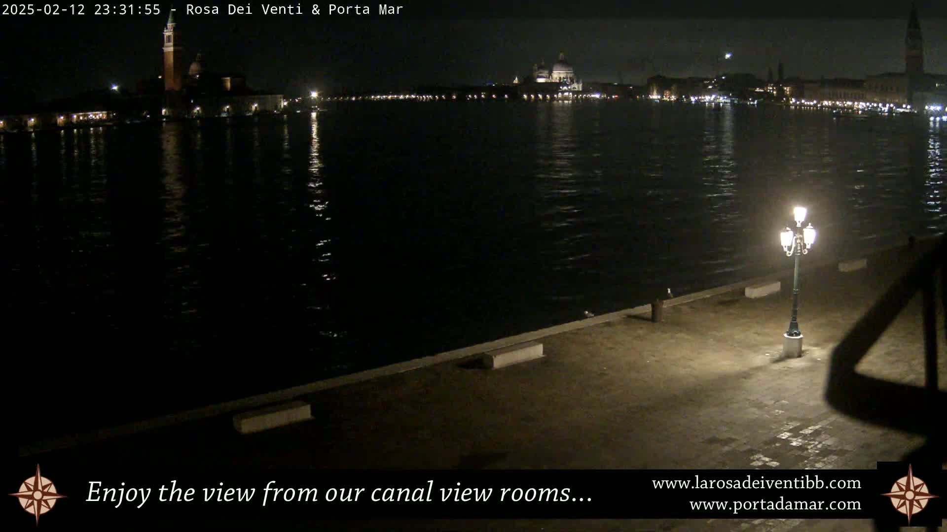 A nighttime view across a calm canal shows illuminated buildings on the far shore, with a single lamppost illuminating a paved area in the foreground.