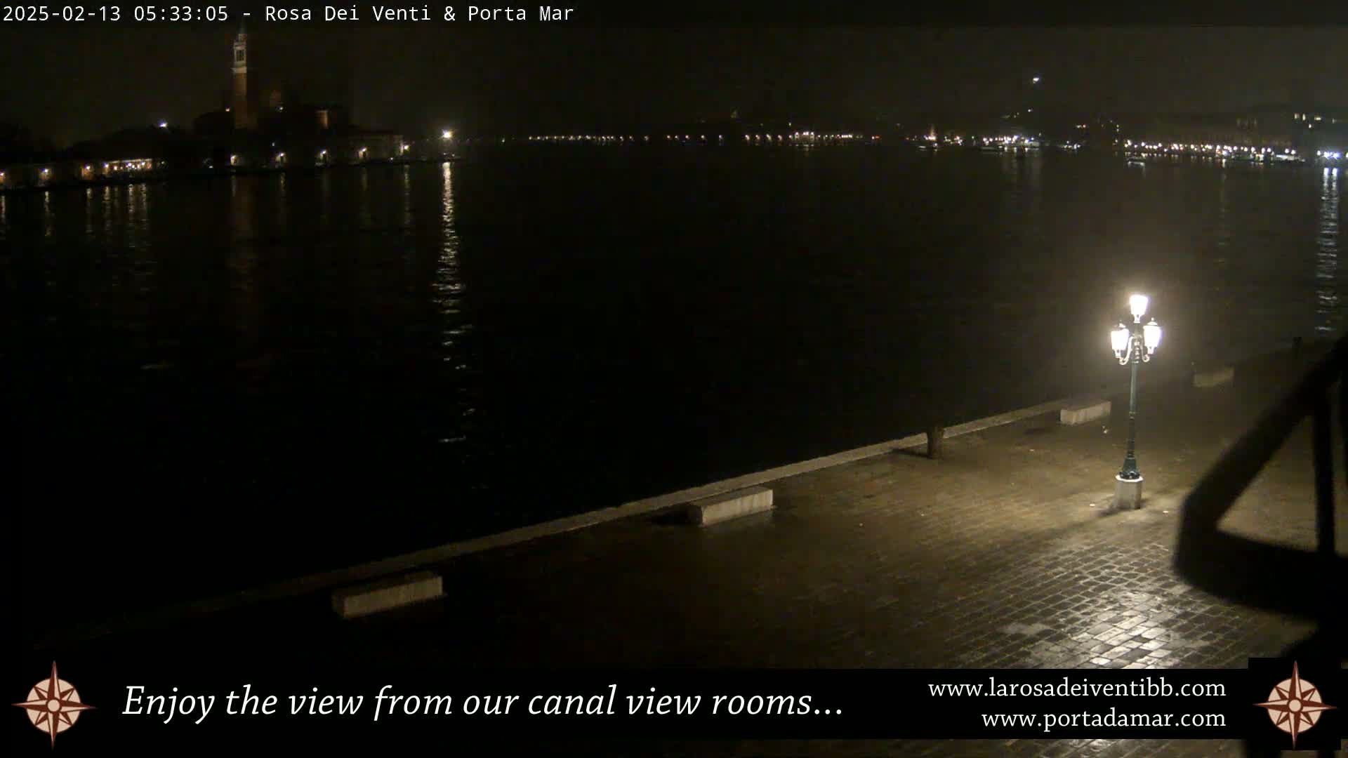 A nighttime view of a calm canal, reflecting city lights, with a single lit lamppost on a cobblestone embankment.