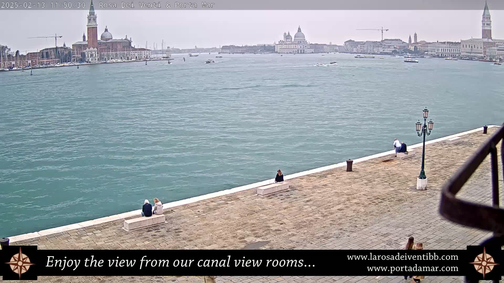 A wide shot of a calm, teal-colored canal in Venice, Italy, shows several people sitting on a stone embankment with buildings and churches in the background on an overcast day.