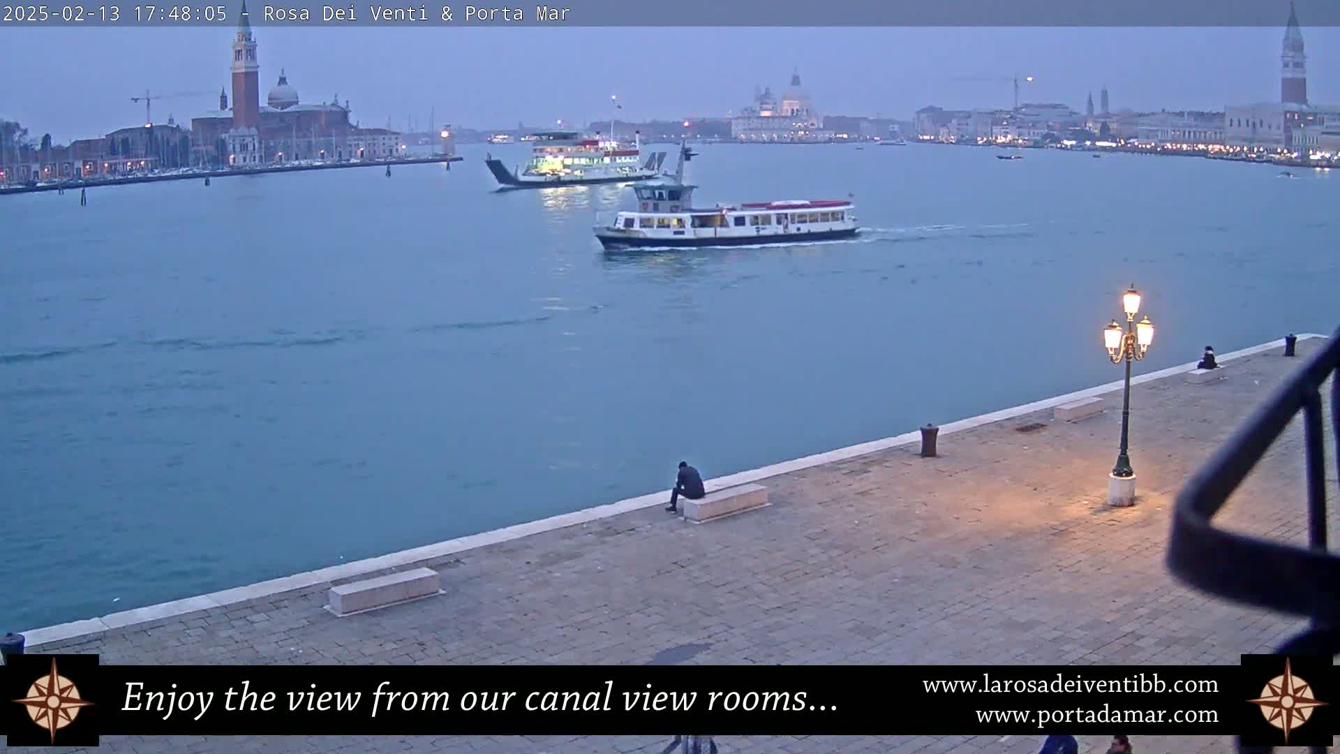 A person sits on a low wall overlooking a calm canal where two ferries are passing by, with a city skyline including a bell tower in the background under a twilight sky.