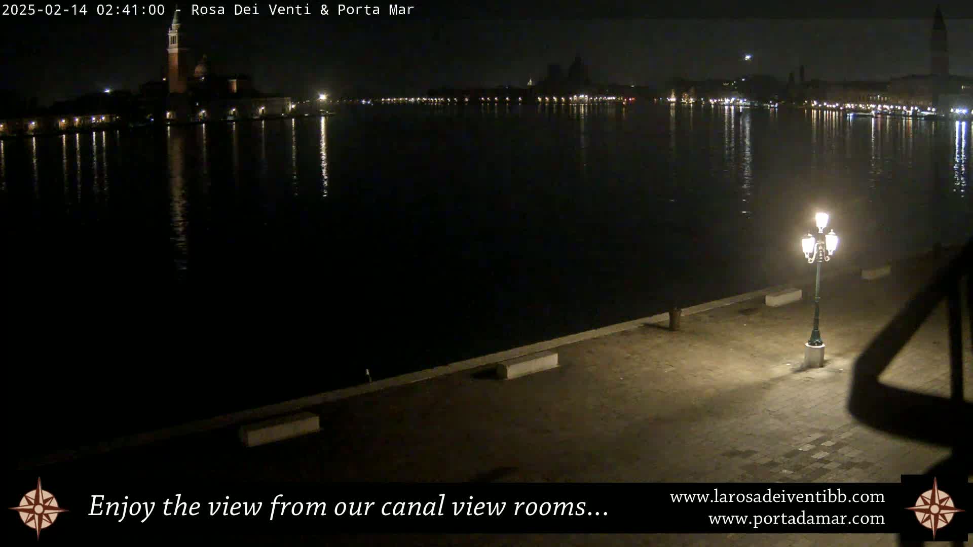 A nighttime view across a calm body of water shows a city skyline with lights reflecting on the water, and a single lit lamppost on a paved waterfront.