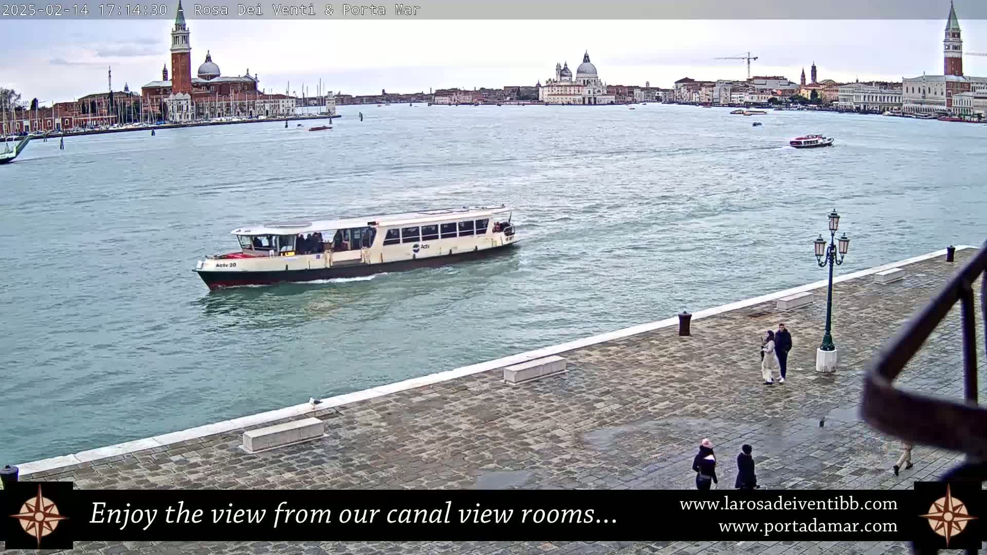 A water taxi travels on a canal toward a city with large buildings and a domed church visible in the distance under an overcast sky.