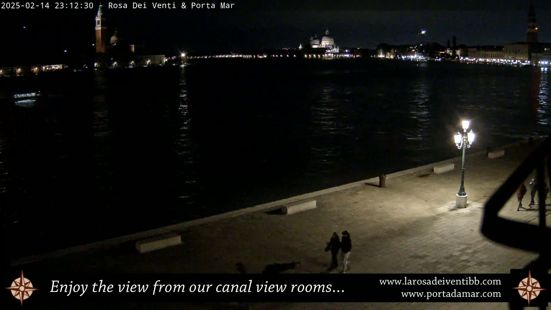 A nighttime view of a calm canal, illuminated by a single lamppost, shows a cityscape across the water with a large domed building, and two people walking along the canal's edge.