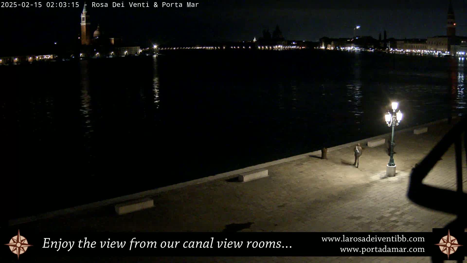 A nighttime view across a calm canal shows a city skyline with lights, a single person walking on a paved waterfront, and a lit streetlamp.