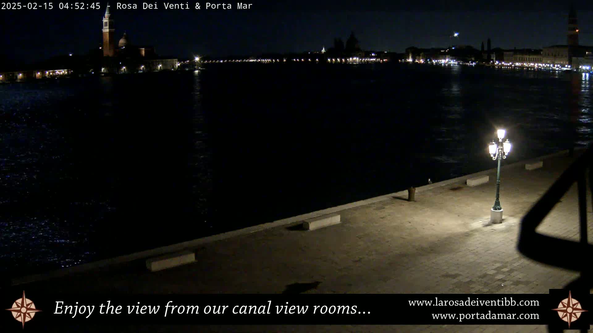 A nighttime view across a dark canal shows a city skyline illuminated with lights, a single lamppost on a paved waterfront, and a dark, calm body of water.