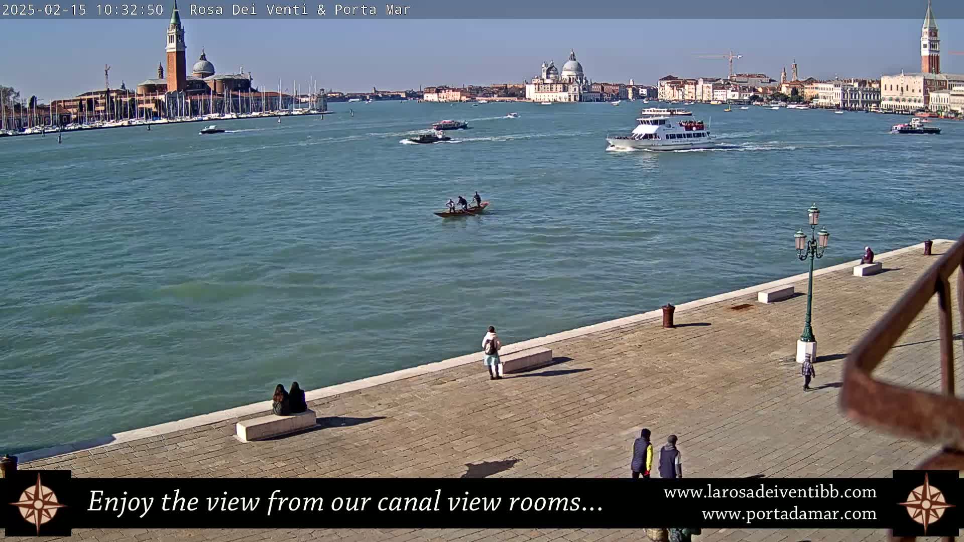 A sunny day in Venice shows a waterway with several boats, including a gondola and a passenger ferry, with the city's buildings and churches visible in the distance.