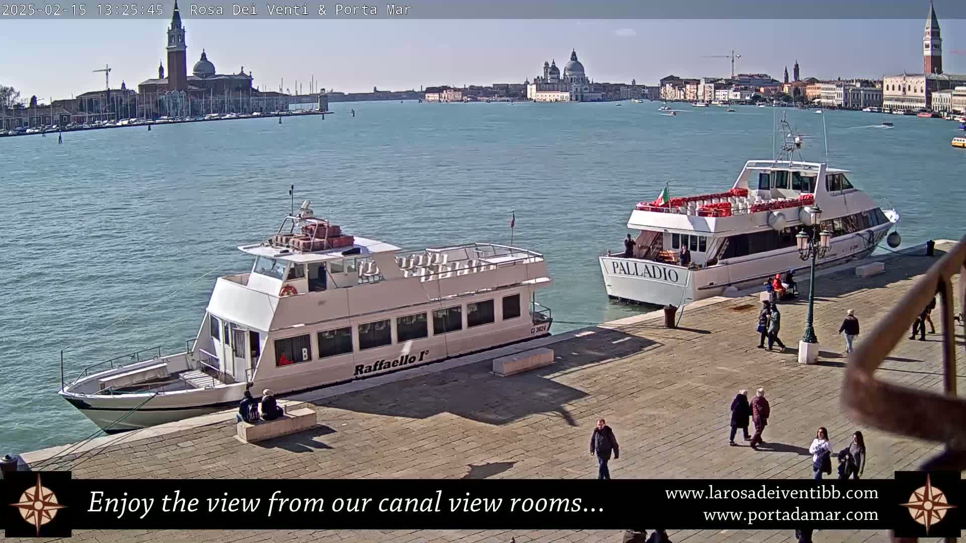 Two passenger boats are docked near a waterfront paved area with pedestrians, overlooking a calm body of water with a distant cityscape under a sunny sky.