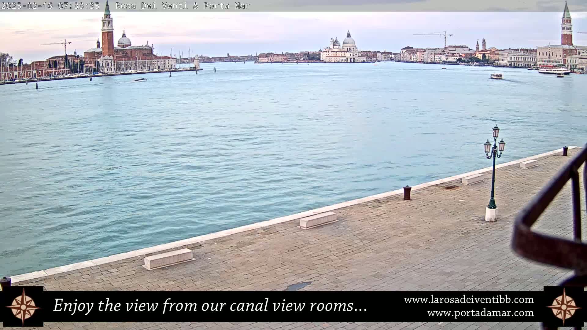 A calm, light-blue canal stretches out to a distant cityscape featuring several prominent churches under an overcast sky.