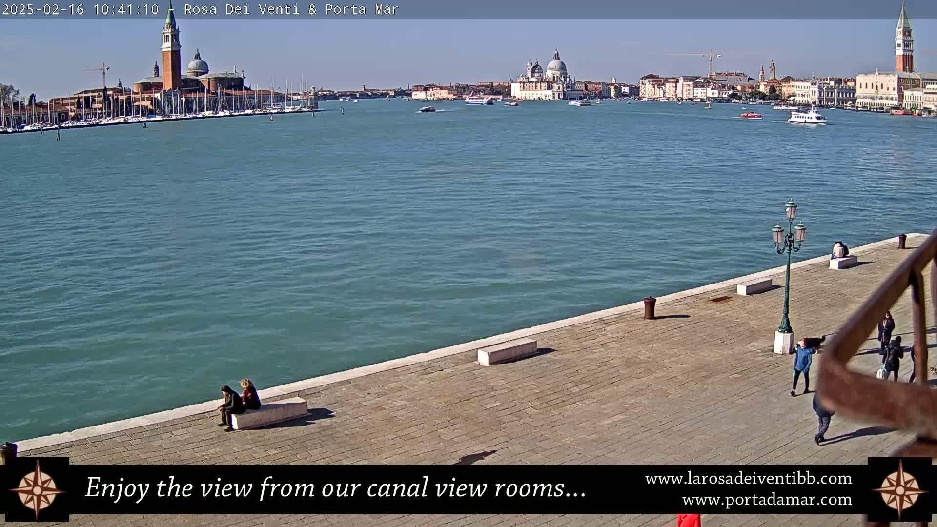 A sunny day in Venice shows a calm canal with boats and a view of the city's buildings, including a prominent church with domes, all seen from a paved waterfront area with a few people walking and sitting.