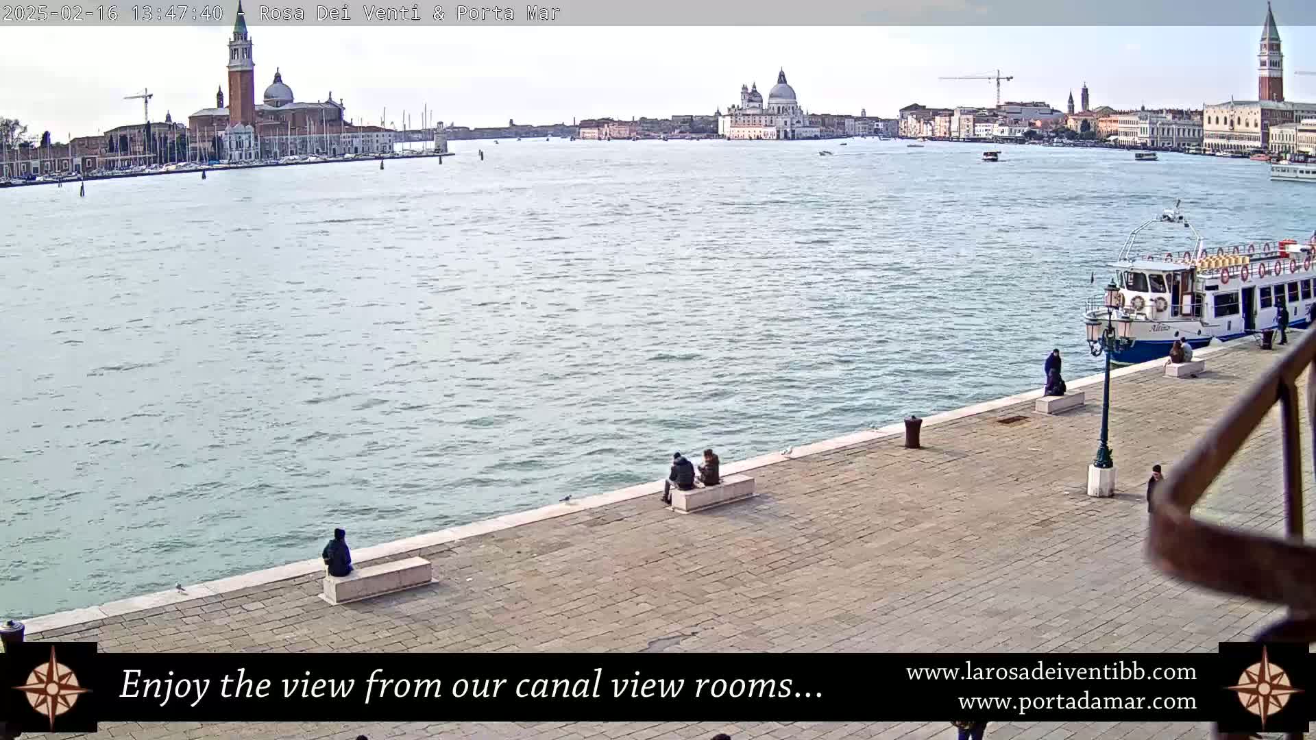 A calm canal with a distant cityscape under an overcast sky, viewed from a paved waterfront where several people sit.