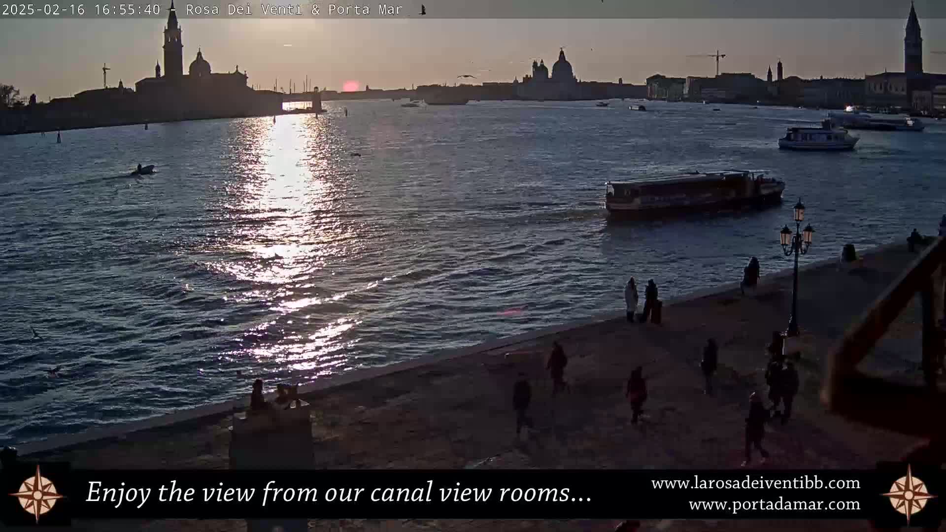 A waterway reflecting the setting sun, with several boats and people walking along a stone embankment, and a city skyline in the background.