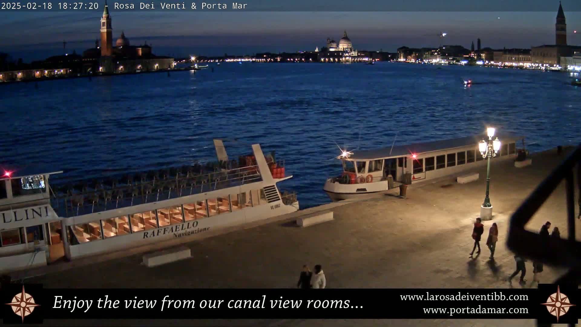 Two passenger boats are docked at a waterfront at night, with a city skyline visible across a dark body of water under a clear, twilight sky.