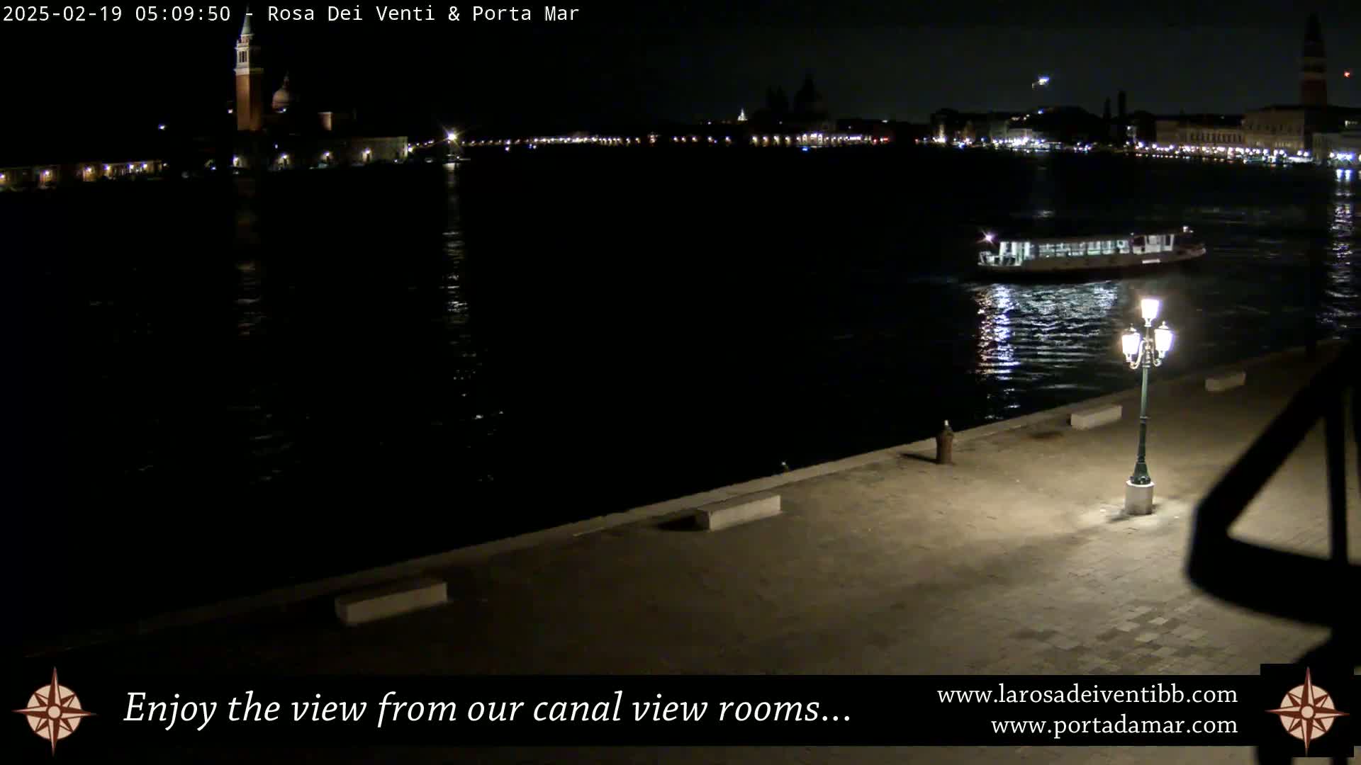 A nighttime view of a calm canal shows a distant illuminated cityscape and a single boat, with a lit lamppost on a paved embankment in the foreground.