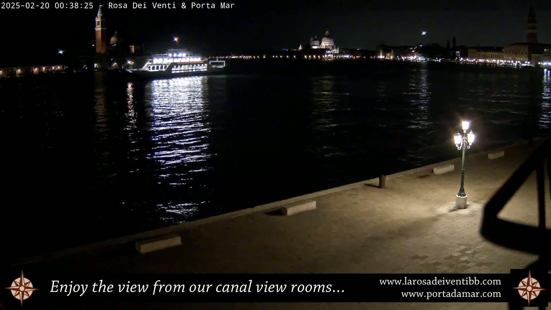 A nighttime view across a dark canal shows a boat and city lights reflecting on the water, with a single lamppost on a paved area in the foreground.