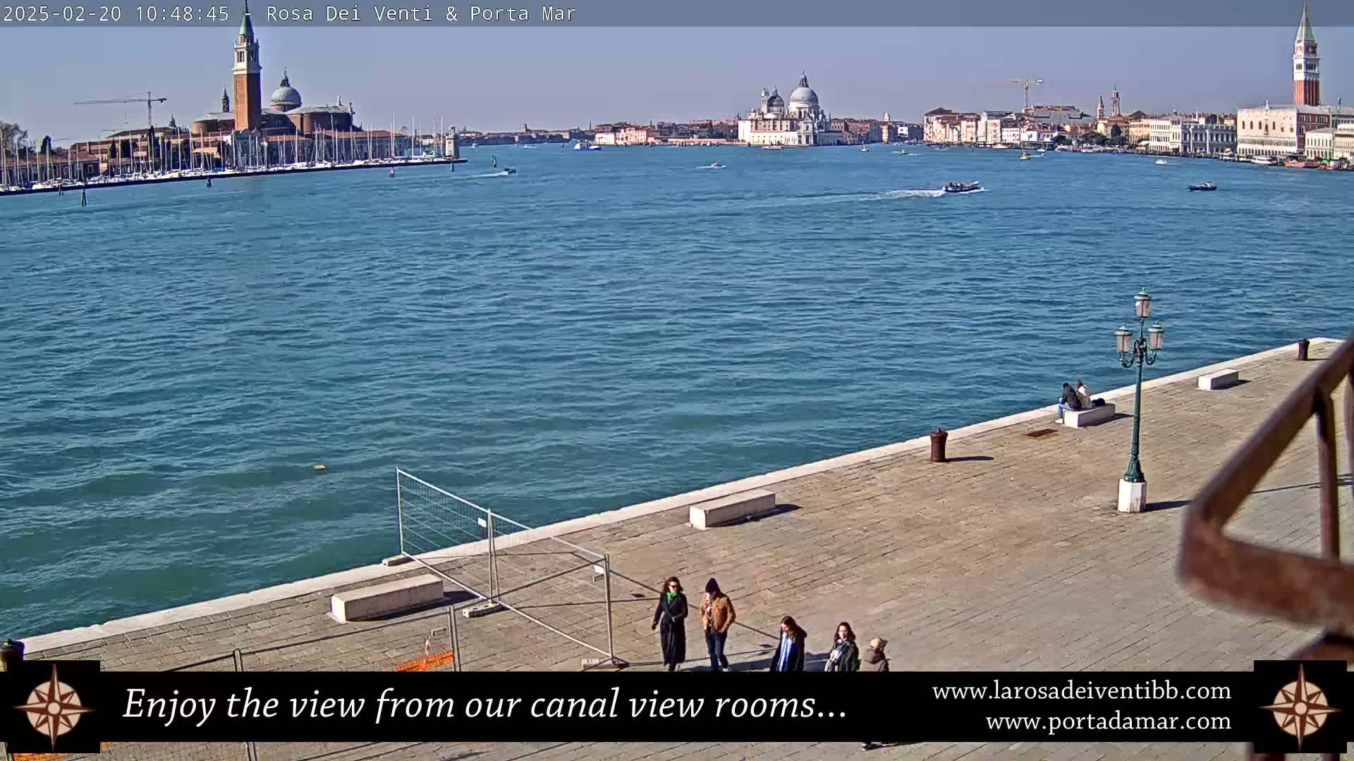 A sunny day in Venice shows a waterfront paved area with a few people, a view of the canal, and buildings in the distance.