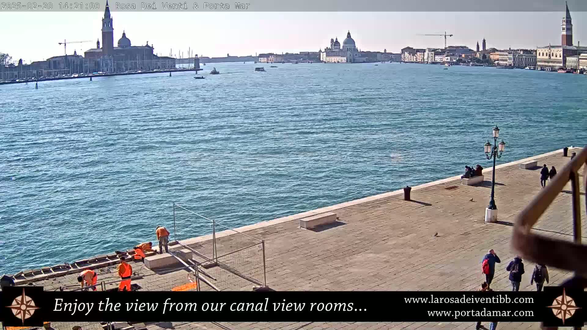 A sunny view of a canal in Venice, Italy, shows workers on a paved walkway near the water, with several buildings and a church visible across the water.