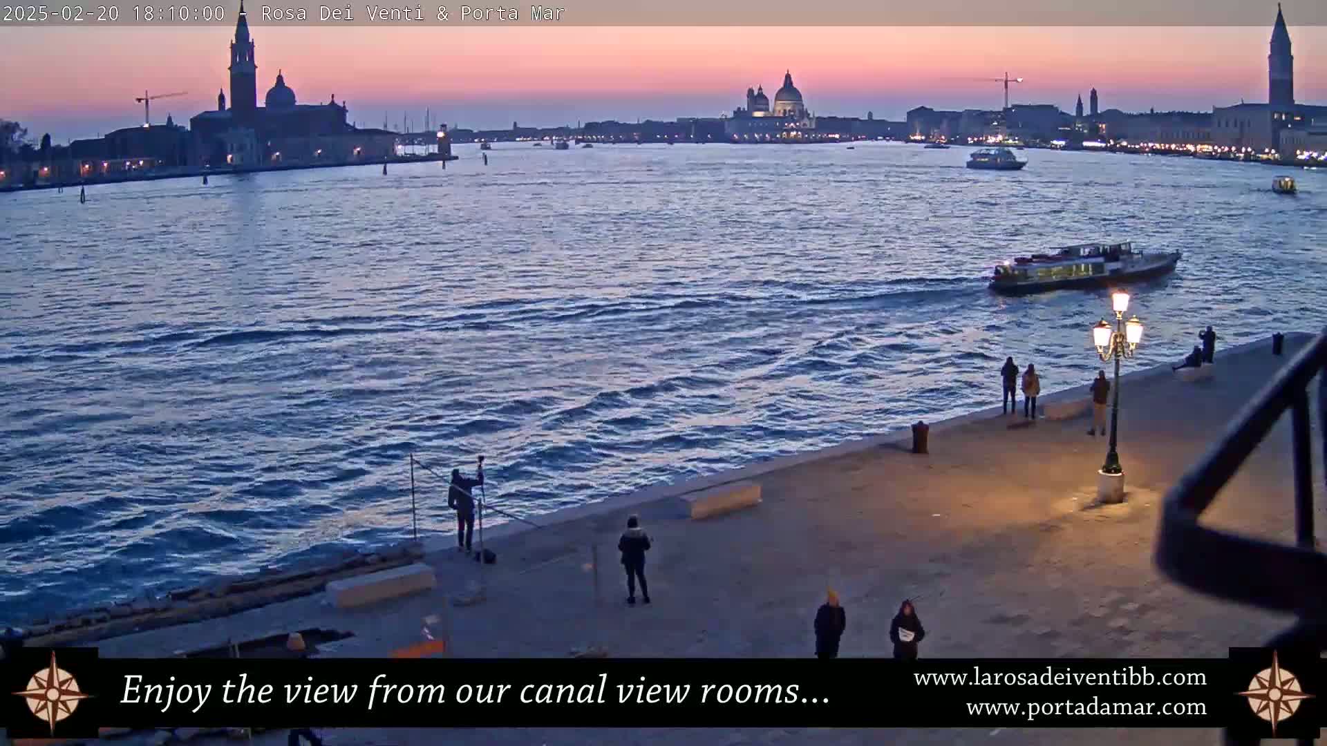 A twilight view over a calm canal shows a distant cityscape with several prominent domed buildings, a few small boats on the water, and several people standing on a paved walkway.