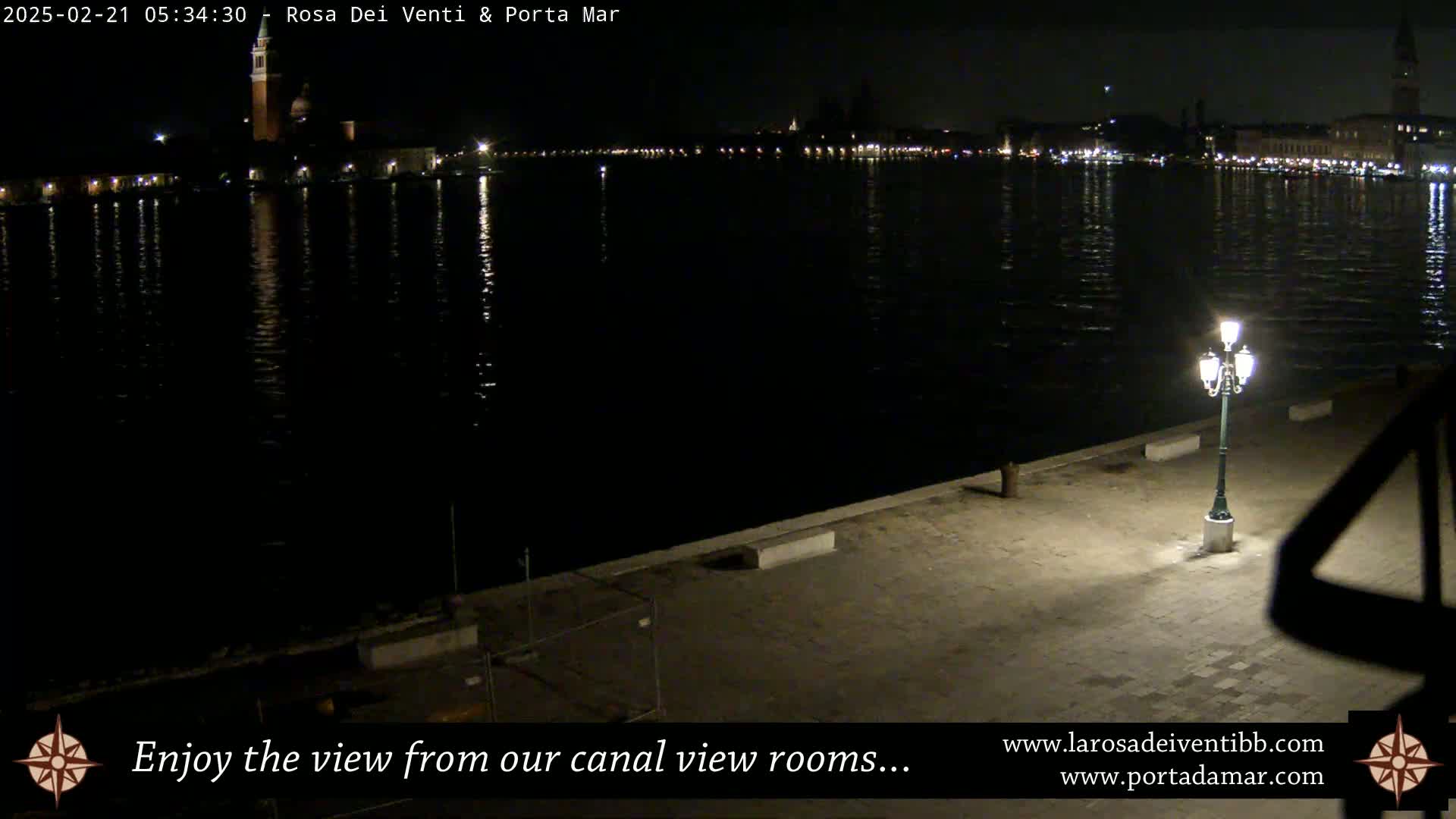 A nighttime view of a calm body of water reflecting city lights across a dark waterfront, illuminated by a single lamppost.