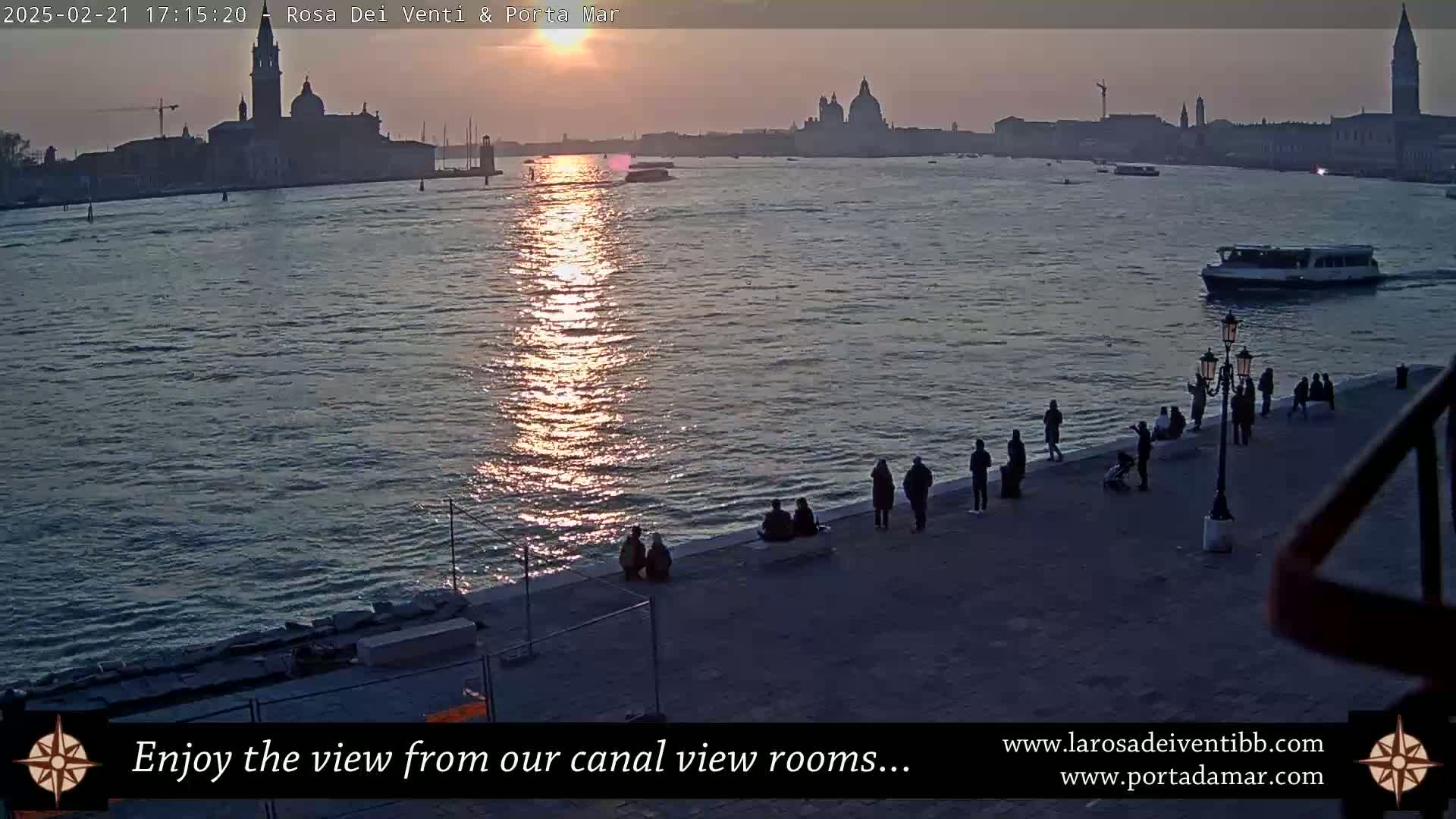 A sunset over a calm body of water shows a city skyline in the distance with several people standing along a waterfront walkway, a boat passing by.
