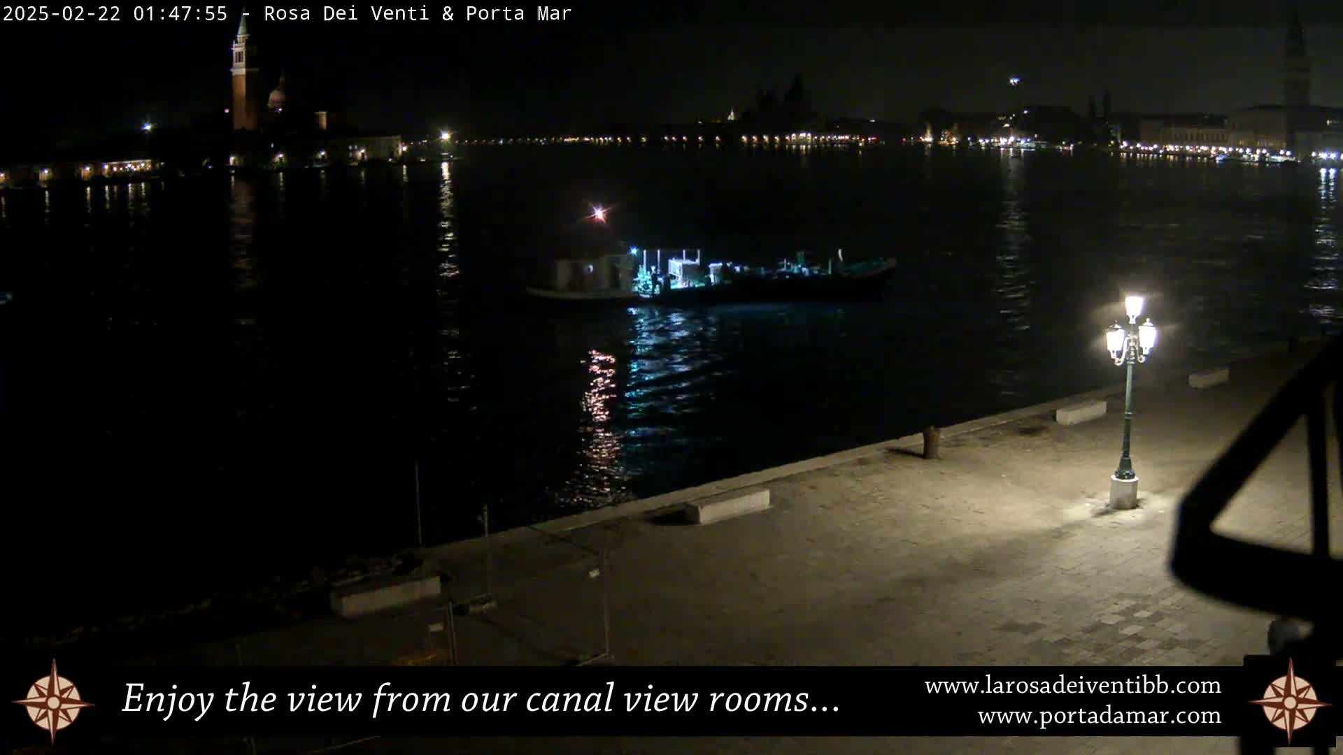 A nighttime view of a canal with a boat and city lights across the water, illuminated by a single lamp on a stone embankment.