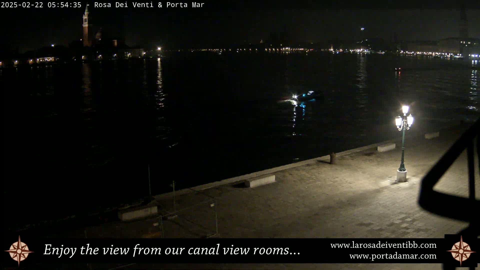 A nighttime view of a calm canal, with a single lit lamppost on a stone embankment, and distant city lights across the water.