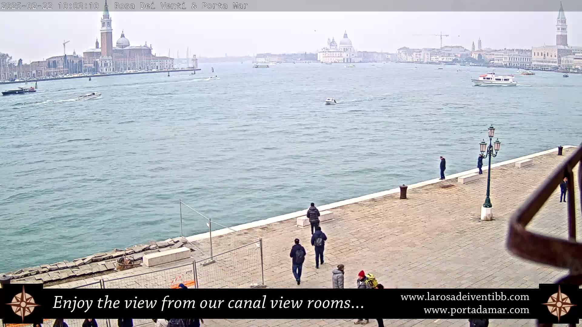 A wide, calm canal in Venice, Italy, on an overcast day, is viewed from a waterfront walkway with several people strolling along it, showcasing buildings and a church in the distance.